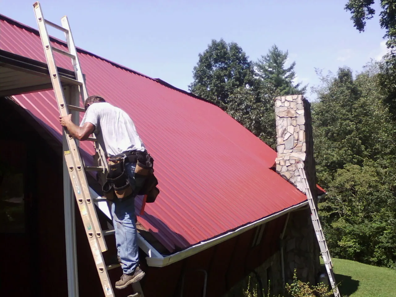 Roofer on a ladder beside a red metal roof and stone chimney — Soffit & Fascia Repair by Roofing Brothers in Maxwell